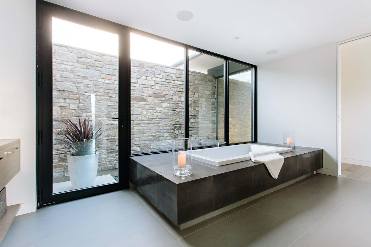 Spa-style bathroom with floor-to-ceiling glass windows, warm stone surfaces, and generous natural light flooding an open accessible layout.