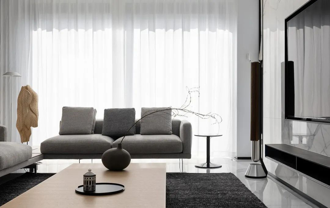 Minimalist living room with a gray sofa in front of sheer floor-to-ceiling curtains, a marble TV wall, and a wood sculpture.