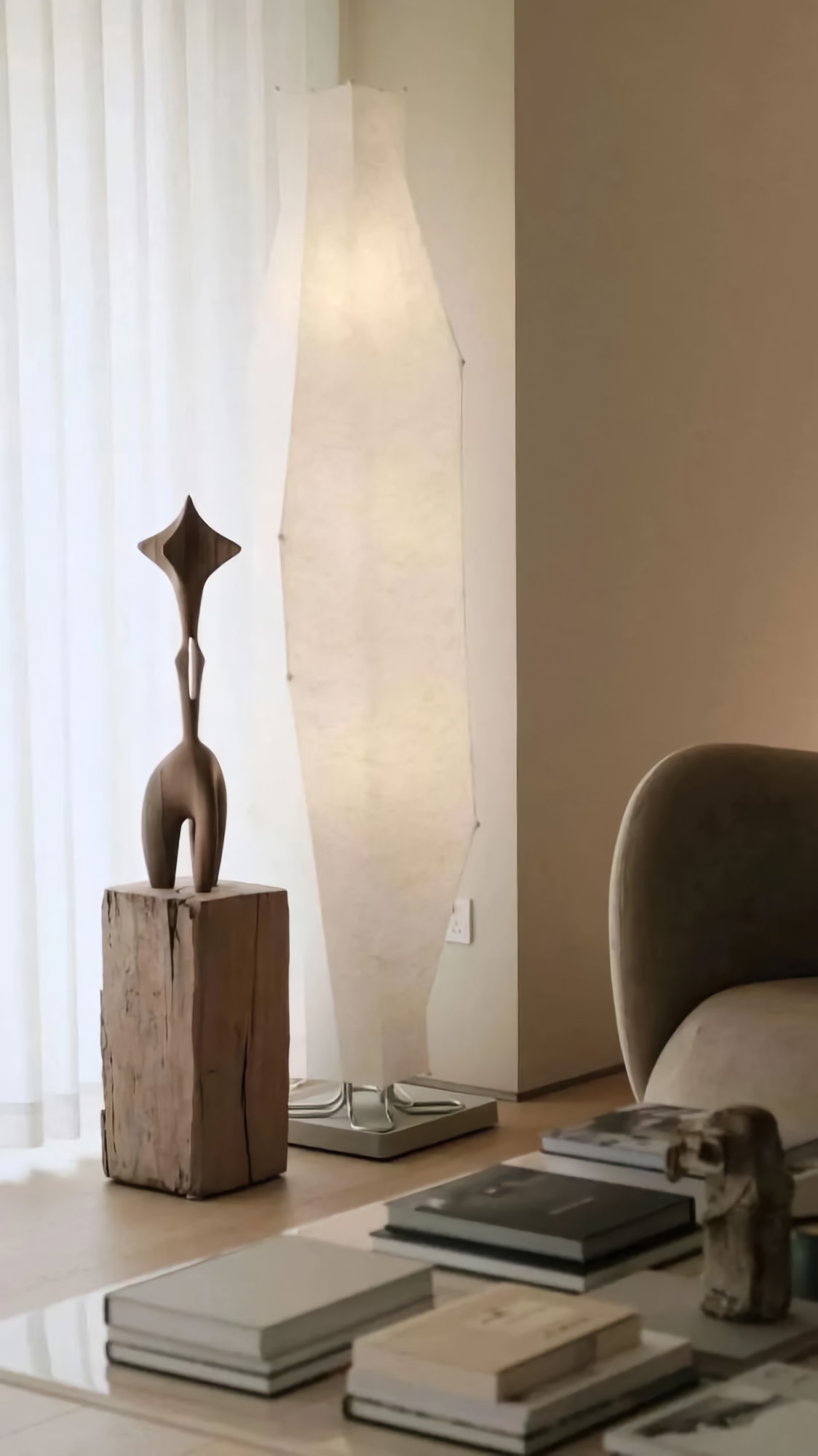 Minimalist living room corner with a warm fabric floor lamp glowing beside a wood sculpture and stacked books.