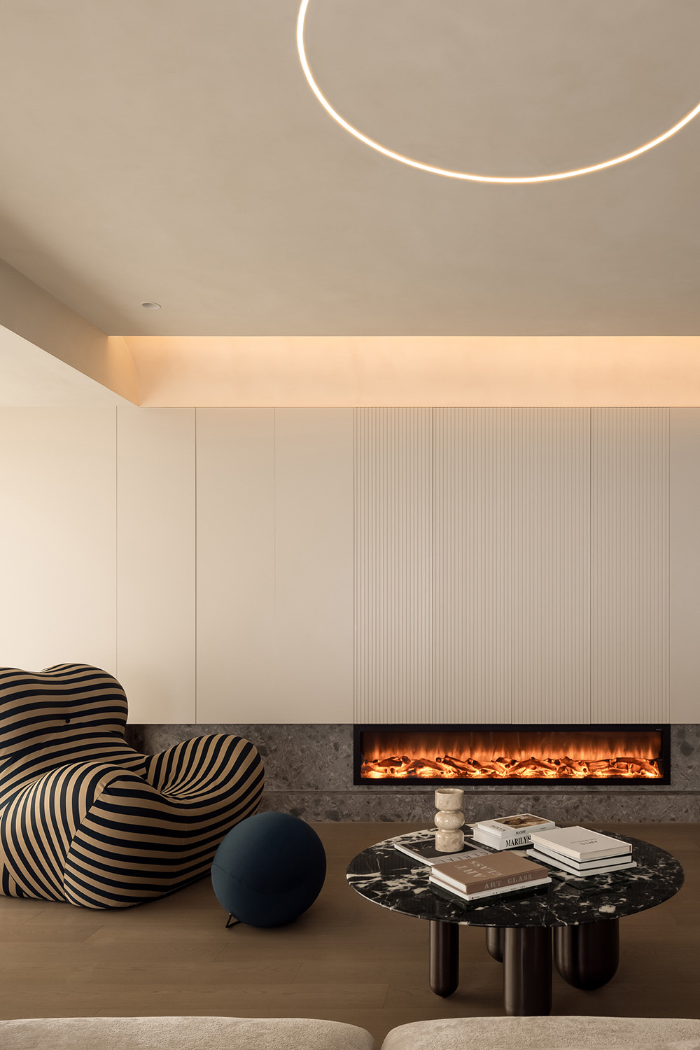 Warm minimalist living room with a glowing ring pendant, ceiling cove lighting, and a lit fireplace insert.