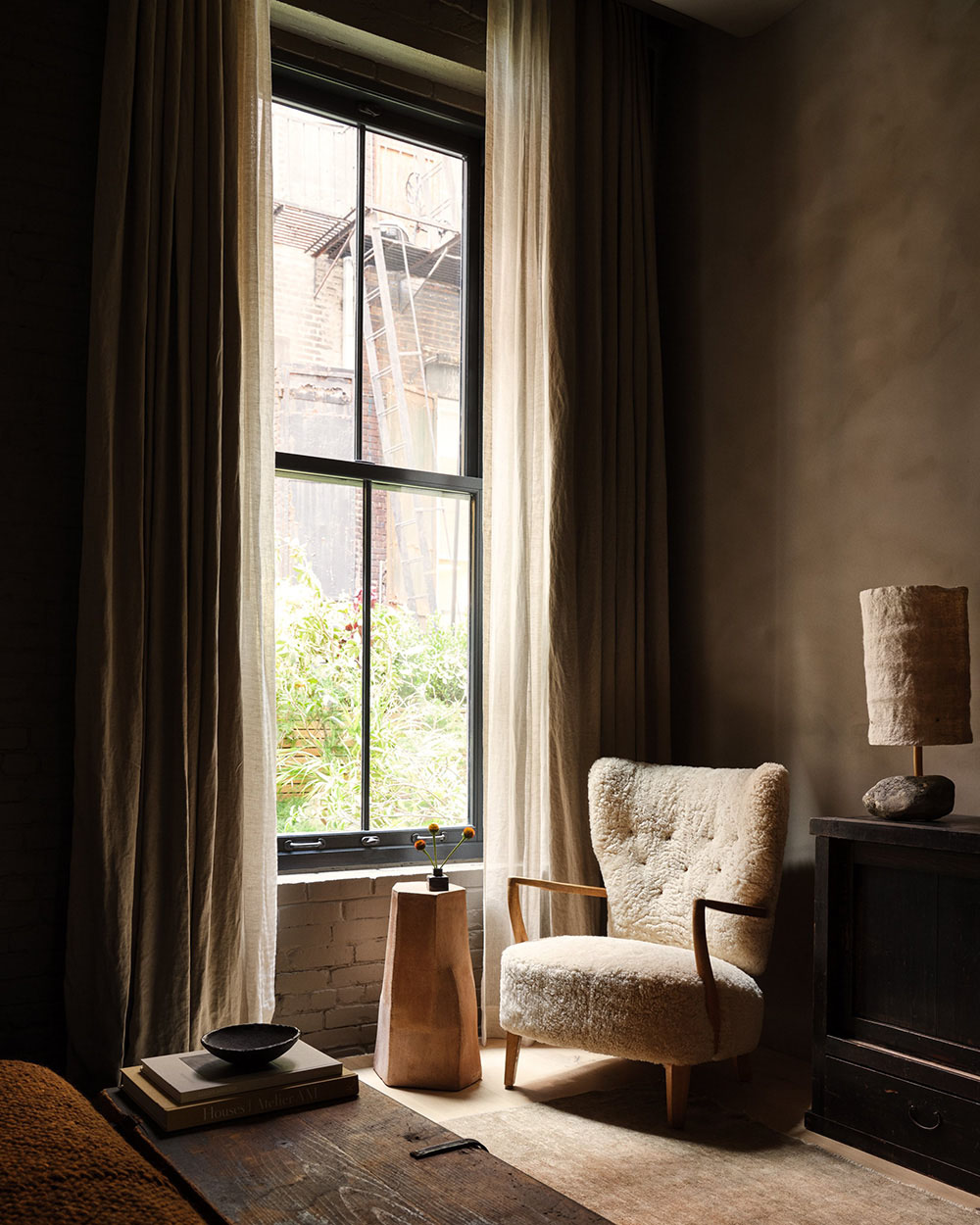 Shearling wingback armchair with wood armrests beside a curtained window and a stack of books on a stool.