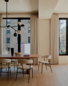 A modern dining room featuring a round oak table surrounded by upholstered chairs, large windows with curtains, and a chandel