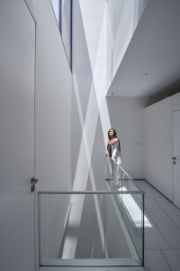 A woman stands on a glass wall railing in a modern, white interior space.