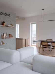 A minimalist living room with concrete walls, a white sofa, and a dining area with a black table and bamboo chairs.