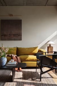 A minimalist living room with a mustard yellow velvet sofa, black chairs, and a wooden lamp on a wooden table with a vase of 