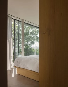 A minimalist bedroom featuring a wooden bed platform and large windows looking out to greenery.