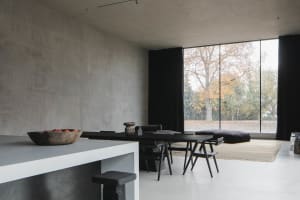 A modern living room with concrete walls and floor, featuring a large window with black curtains, a black wooden dining table