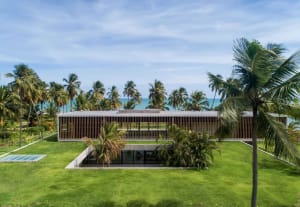 A modern house with a concrete exterior surrounded by lush greenery and palm trees, overlooking the ocean.