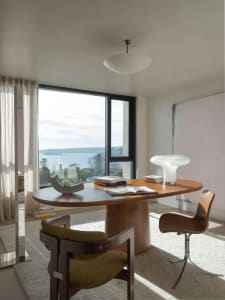 A modern residential office space featuring a wood desk and chairs, with books and a lamp on the desk, and a large window ove