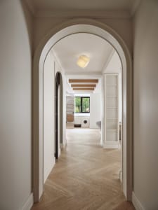 A modern hallway with white walls, wooden flooring, and a view through an archway to a lounge area with white furniture and l