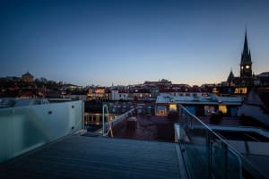 A rooftop view of a cityscape at dusk, featuring illuminated windows and a steeple in the background.