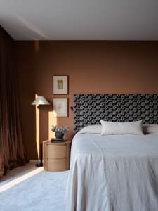 A modern bedroom with a patterned upholstered headboard, white bedding, a wooden bedside table, a floor lamp, and a framed ar