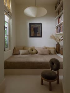 A minimalist bedroom featuring a textile sofa with round cushions, a hanging light fixture, and a bookshelf filled with books