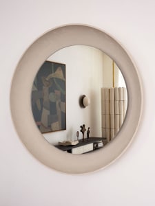 A modern circle wall mirror with a textured concrete frame hanging on a light-colored wall, reflecting a living room with a w