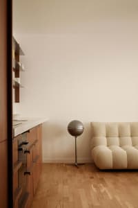 A minimalist living room featuring a beige tufted sofa, a spherical floor lamp, and wooden cabinets against a plain wall on a