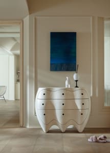 A modern, white dresser with a sleek, organic shape and a blue abstract painting above it in a well-lit living space.