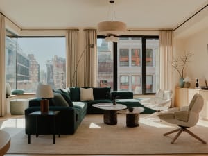 Modern living room with a green sectional sofa, white chairs, and a large window overlooking the city.