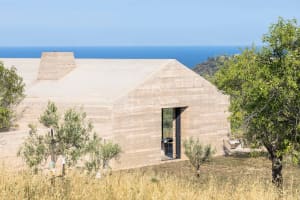 A modern residential house with concrete walls and a large glass door, set against a backdrop of greenery and the sea.