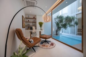 Modern living room with a glass wall overlooking a poolside area, featuring a brown leather chair, ottoman, and circular rug 