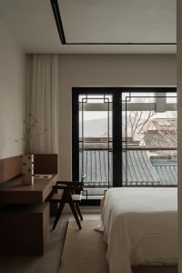 A modern minimalist bedroom with a white bed, wooden furniture, and a view of tiled rooftops.