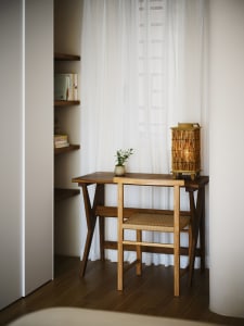 A minimalist home office with a wooden desk, chair, and a lantern lamp, featuring sheer curtains and a bookshelf.