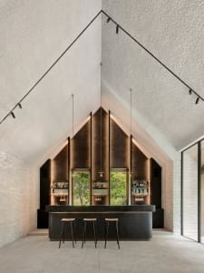 A modern bar interior featuring a concrete and brass counter with three stools, set against a textured wall and glass wall.