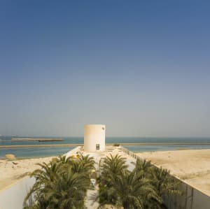 A modern, cylindrical clay tower amidst tropical palm trees by the waterfront.