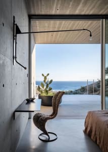 A modern bedroom featuring a wicker chair and a cactus plant in front of a concrete wall overlooking the sea.