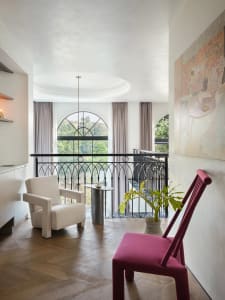 A modern living room with wooden furniture, a white armchair, a pink chair, and a large arched window.