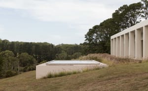 A minimalist house with a concrete exterior and glass windows, surrounded by a grassy landscape.
