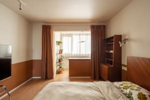 Modern bedroom with wooden furniture and curtains, featuring a bed with a floral comforter, a bookshelf, and a window with pl