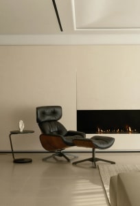 A minimalist living room featuring a black leather and wood armchair with a footstool, a side table with a sculpture, and a m