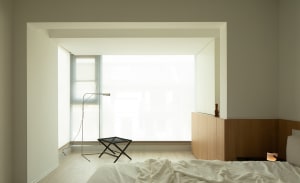 A modern minimalist bedroom with a large window, natural light, a bed, and a black ottoman.
