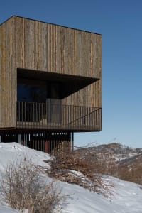 A modern wooden house with a large open window and balcony overlooking a snowy landscape.