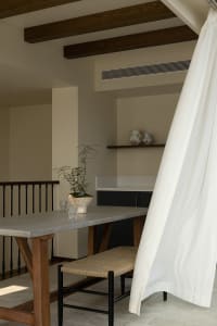 A minimalist industrial-style kitchen with exposed wood beams, concrete countertops, and a plant on the table.
