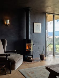 A cozy industrial-style living room features a concrete fireplace, wood floors, and a rustic rug, with a view overlooking a g