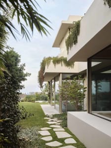 A modern residential garden features a path with stepping stones leading to a house with concrete walls, greenery on the balc