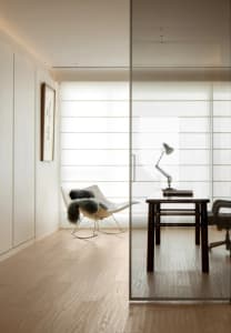 A modern home office featuring a glass wall, wooden desk, and comfortable rocking chair.