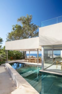 Modern residential exterior featuring a glass wall and concrete patio with a swimming pool and dining area.