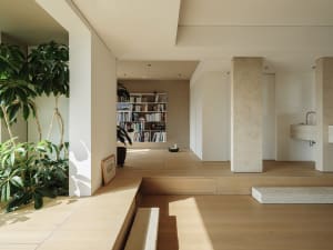 A modern living room featuring wooden flooring and concrete structural columns, with a bookshelf filled with books and potted