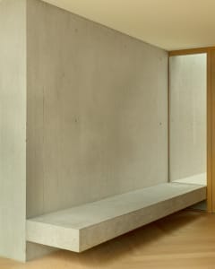 A minimalist interior with a concrete floating shelf against a concrete wall on a light-colored hardwood floor.