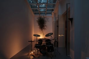 A room with an industrial-style design featuring a drum set, hanging plant, and concrete floor, illuminated by a candle.
