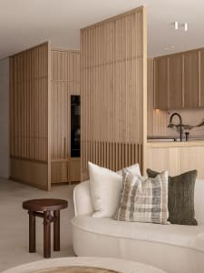 A modern residential interior featuring a white sofa with patterned and solid pillows, wooden furniture, and a kitchen area w