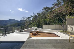 A modern roof deck features a curvilinear concrete seating area with green cushions, a wooden fire pit table, and a view of t