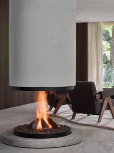 A modern living room with a circular concrete fireplace, wooden furniture, and a view of greenery outside.
