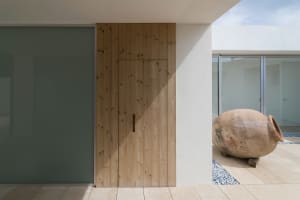 A minimalist wooden door with a handle, set against a concrete exterior wall with a large vase on the floor.