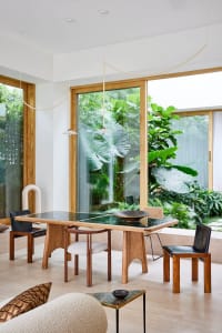 A modern dining room featuring a wooden table with green tiles, surrounded by natural light and lush greenery outside the win