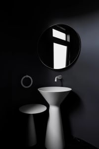 A modern bathroom space with black walls, a metallic pedestal sink, and a round mirror hanging above.