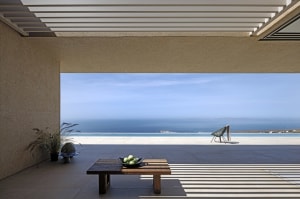 A modern outdoor patio with concrete walls and floor, featuring a wooden table, green plants, and a view of the ocean.