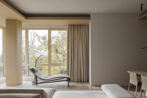 A minimalist living room with neutral fabrics and wood elements, featuring a lounge chair, sofa, and large windows with a vie